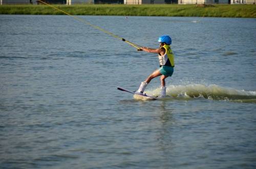 A child wearing a helmet and life jacket is water skiing on a calm lake, creating a small splash behind them.