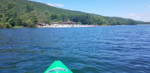 A view from a kayak on a calm lake, with a beach and green hills in the background under a clear blue sky.