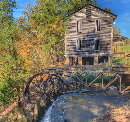 A rustic wooden mill with a waterwheel, surrounded by trees and autumn foliage. Water flows from the wheel.