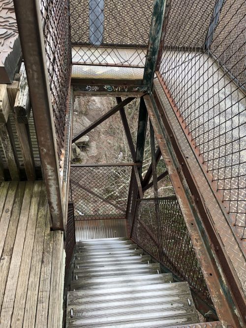 A view looking down a metal staircase with a wooden landing, surrounded by a mesh fence and a natural landscape below.