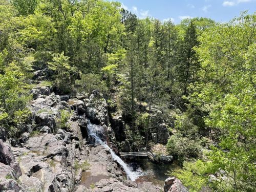 A serene landscape featuring a small waterfall surrounded by lush green trees and rocky terrain under a clear blue sky.