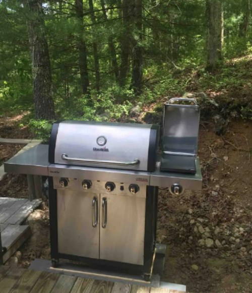 A stainless steel gas grill on a wooden deck, surrounded by trees in a natural outdoor setting.