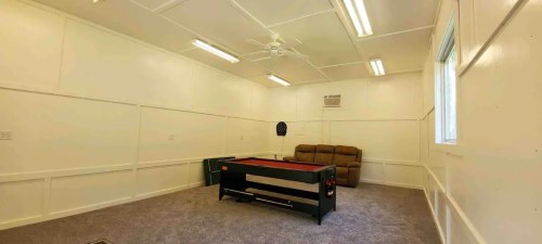 A spacious, empty room with a pool table, a brown couch, and white walls, featuring overhead lights and a ceiling fan.