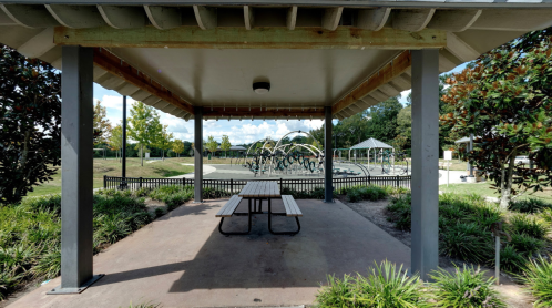 A shaded pavilion with a picnic table overlooks a park with green grass and trees in the background.