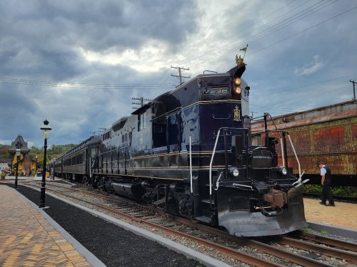 A vintage black train with a gold emblem parked at a station under a cloudy sky, surrounded by tracks and old train cars.