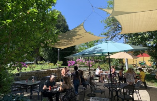 Outdoor dining area with shaded tables, people enjoying meals, and vibrant greenery and flowers in a sunny setting.