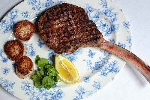 A grilled tomahawk steak on a decorative plate, accompanied by scallops, lemon, and fresh greens.
