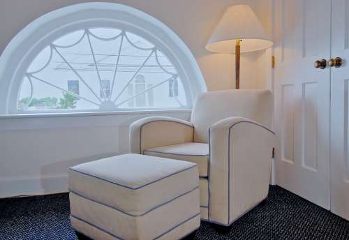Cozy corner with a white armchair and ottoman beside a lamp, featuring a large arched window with decorative design.