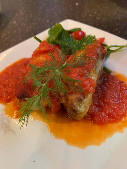 A plated dish featuring a piece of meat topped with red sauce and garnished with fresh dill and greens.