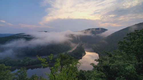 A serene landscape featuring a river winding through mountains, shrouded in mist under a cloudy sky at dawn.