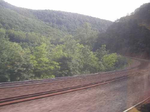 A scenic view of train tracks curving through lush green hills and trees under a bright sky.