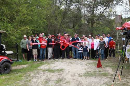 A group of people gathered outdoors, smiling and holding a large red ribbon for a ribbon-cutting ceremony.