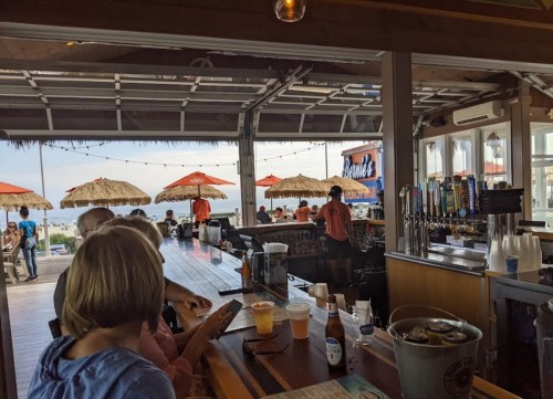 A beachside bar with patrons, umbrellas, and a view of the ocean in the background.