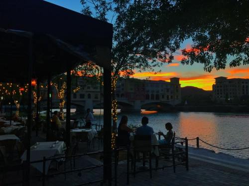 Silhouetted diners at a lakeside restaurant during a vibrant sunset, with colorful skies reflecting on the water.