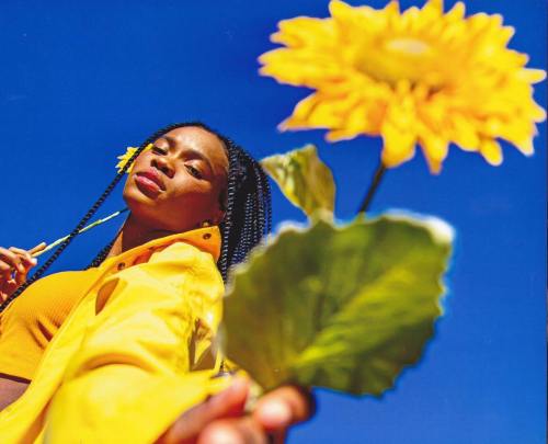 A woman in a yellow jacket holds a sunflower against a bright blue sky, exuding joy and confidence.