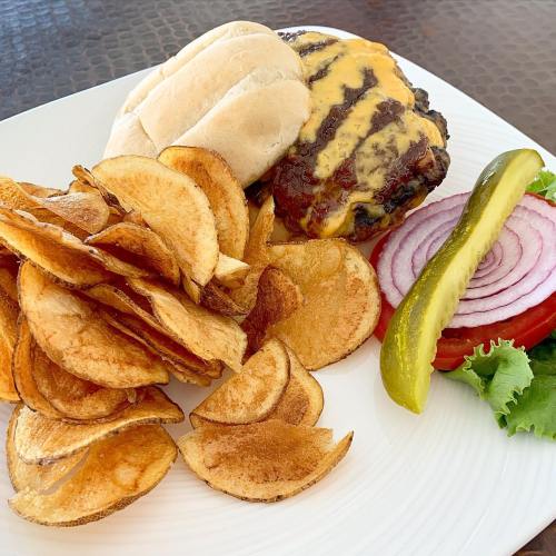 A plate featuring a cheeseburger with a soft bun, served with crispy potato chips, pickles, and onion slices.