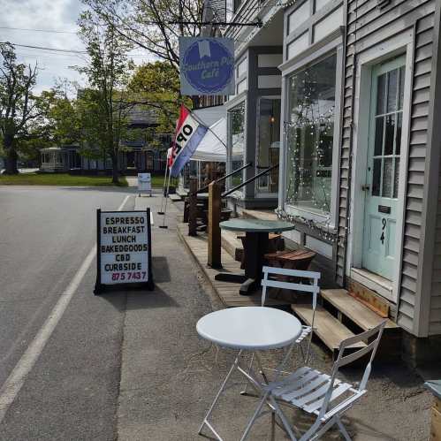 A quaint café exterior with a sign advertising espresso and baked goods, featuring outdoor seating and a sunny street view.