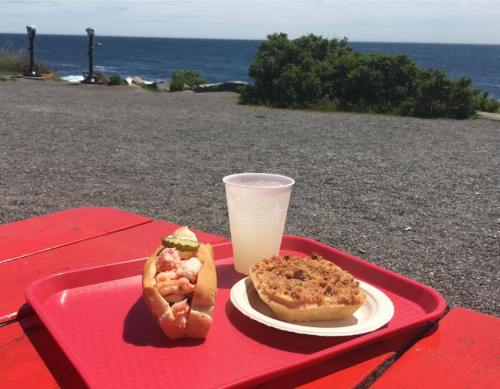 A red tray holds a lobster roll, a dessert, and a cup of lemonade, with a scenic ocean view in the background.