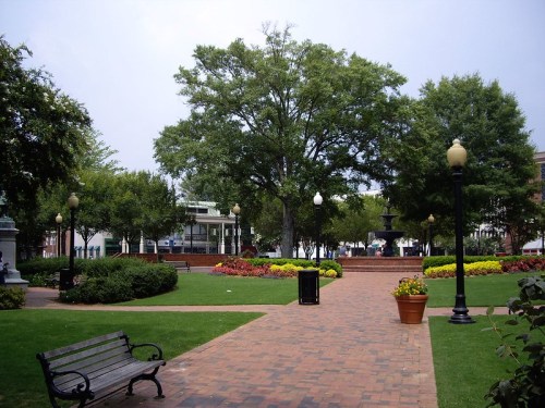 A park with a brick pathway, benches, a fountain, and vibrant flower beds surrounded by trees and lampposts.