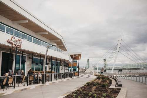 A waterfront restaurant with outdoor seating, alongside a modern bridge and cloudy sky.