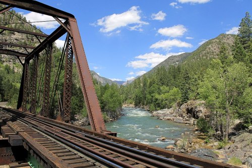 A scenic view of a river flowing beneath a metal bridge, surrounded by lush greenery and mountains under a blue sky.