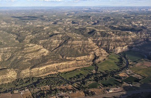 Aerial view of rugged terrain with layered hills, green fields, and a winding river in a vast landscape.