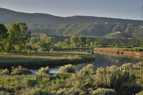A serene river flows through lush greenery, surrounded by rolling hills under a clear sky.