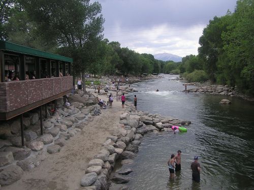 A scenic riverbank with people swimming and relaxing, surrounded by trees and rocky shores, under a cloudy sky.