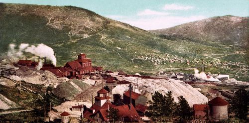 A historic mining landscape with buildings, smokestacks, and hills in the background, showcasing industrial activity.