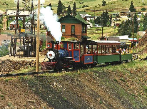 A vintage steam train with a green passenger car travels along a track near a small town and a wooden building.