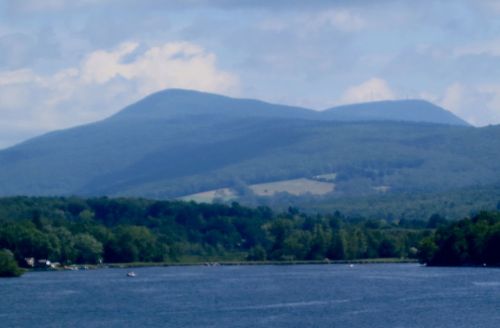 A serene river flows in the foreground, with lush green hills and mountains in the background under a cloudy sky.