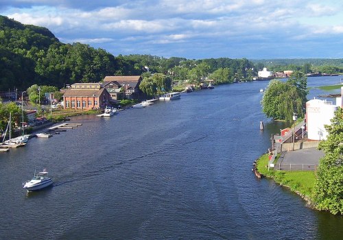 A scenic river view with boats, lush greenery, and buildings along the banks under a partly cloudy sky.