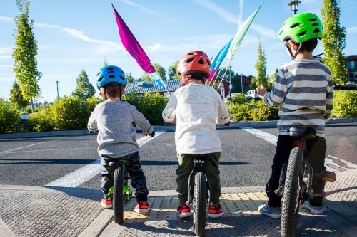 Three children on bicycles, wearing colorful helmets, facing a sunny outdoor scene with colorful flags in the background.
