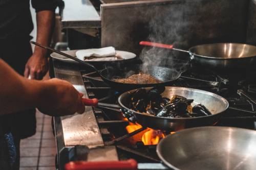 A chef cooks mussels in a pan over an open flame, with another pan sizzling on the stove in a busy kitchen.