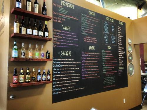 A chalkboard menu featuring breakfast, soups, wraps, salads, and drinks, alongside a shelf of assorted beverages.