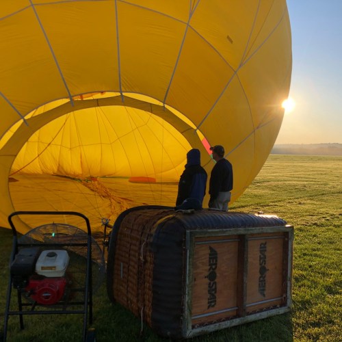 Two people stand inside a yellow hot air balloon, preparing for takeoff on a clear morning.