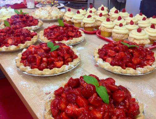 A table filled with strawberry pies and cupcakes topped with cream and raspberries, dusted with powdered sugar.