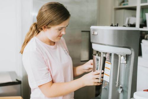 A young woman in a pink shirt uses a silver machine, holding a cup, in a bright kitchen setting.