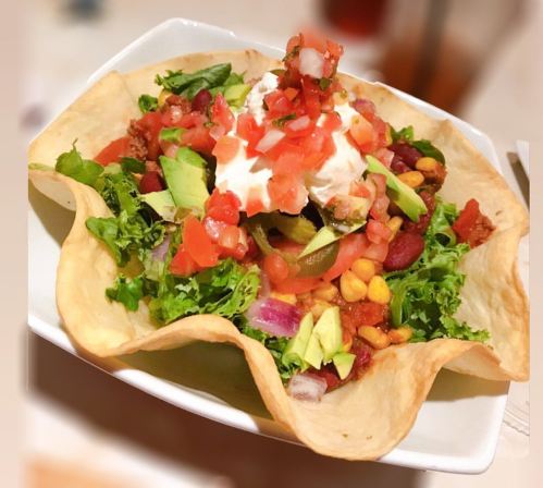 A colorful taco salad in a crispy shell, topped with lettuce, tomatoes, avocado, corn, and sour cream.