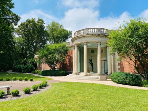 A scenic view of a brick building with columns, surrounded by greenery and a well-maintained lawn on a sunny day.