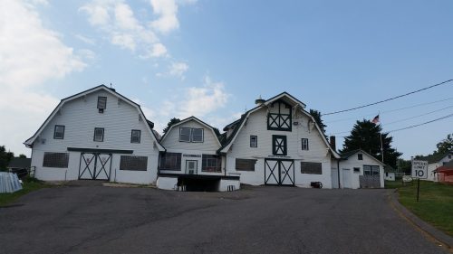 A large white barn with green accents, set on a gravel road, under a cloudy sky. An American flag is visible nearby.