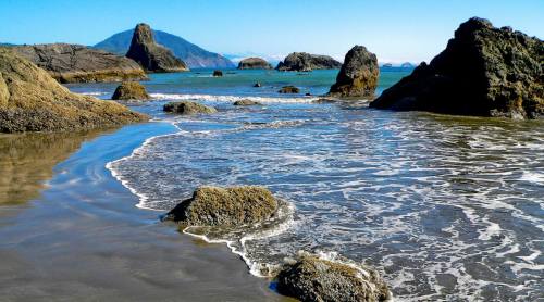 A serene beach scene with rocky formations, gentle waves, and a distant mountain under a clear blue sky.
