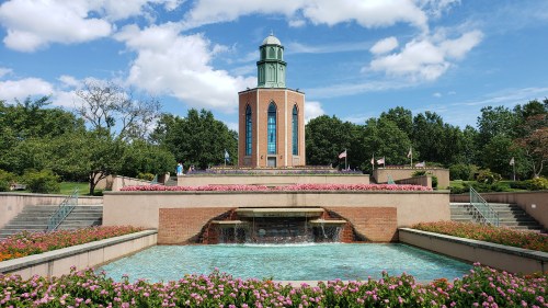 A fountain surrounded by colorful flowers in front of a tall building with a green dome under a blue sky.