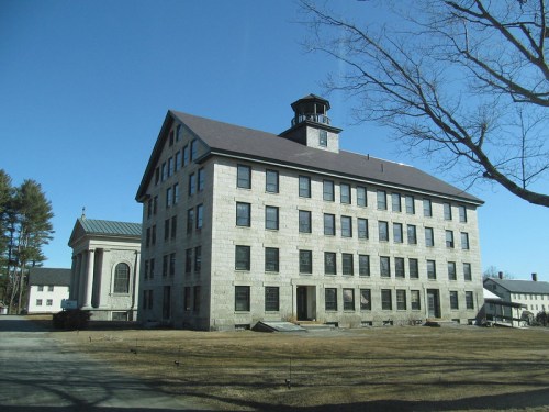Historic stone building with a tower, surrounded by a grassy area and trees, under a clear blue sky.