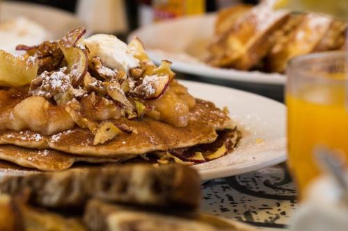 A stack of pancakes topped with apples, nuts, and whipped cream, served with toast and a glass of orange juice.