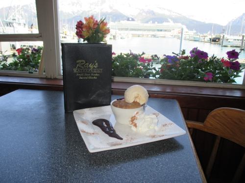 A dessert with ice cream and whipped cream on a plate, next to a menu, with a scenic waterfront view in the background.