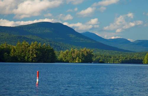 A serene lake surrounded by lush green mountains under a partly cloudy sky, with a red and white buoy in the foreground.