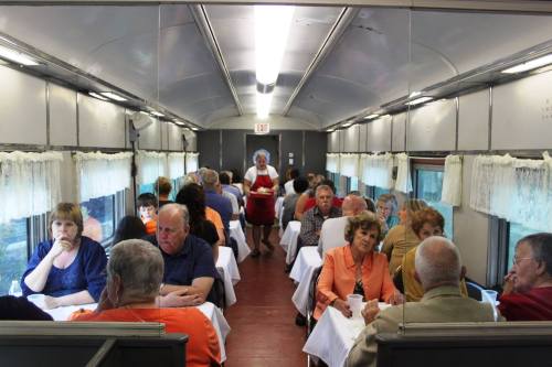 A dining car filled with people at tables, with a waitress serving in the aisle. Brightly lit and cozy atmosphere.