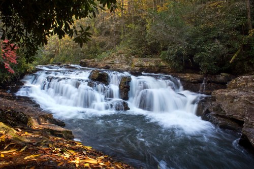 A serene waterfall cascades over rocks, surrounded by autumn foliage and lush greenery.