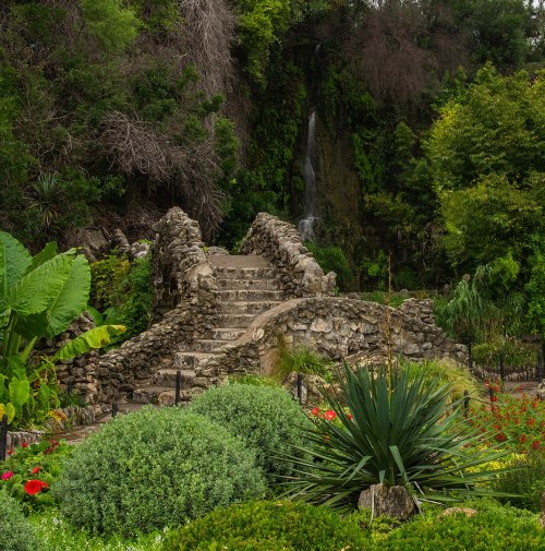 A stone bridge leads to a waterfall, surrounded by lush greenery and colorful flowers in a serene garden setting.